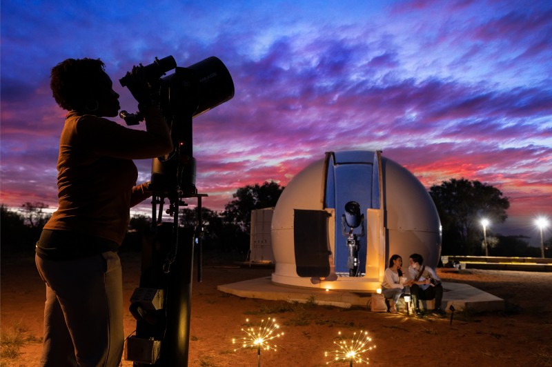 sunset image with woman looking through telescope and couple sitting in front of dome