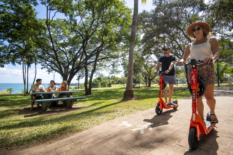 Man and woman on scooters riding along Darwin Esplanade. People enjoying picnic in the background.